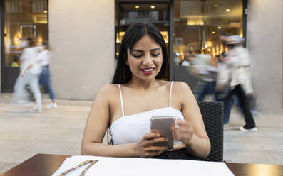Smiling young woman using mobile phone at sidewalk cafe - JCCMF02659