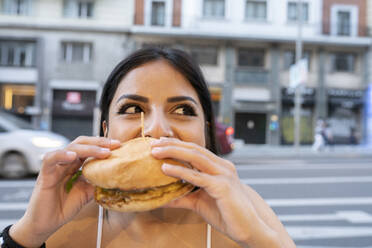 Junge Frau isst einen Hamburger in einem Straßencafé in der Abenddämmerung - JCCMF02664
