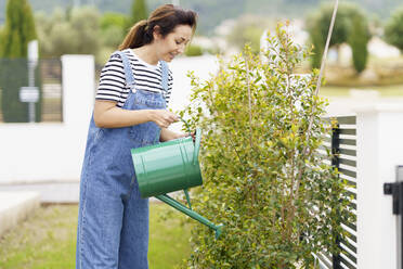 Lächelnde Frau im mittleren Erwachsenenalter gießt Wasser auf Pflanzen im Garten eines neuen Hauses - JSMF02270