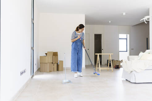 Mid adult woman sweeping living room floor at new home - JSMF02283