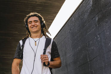 Lächelnder Mann hört Musik über Kopfhörer, während er einen Rucksack an der Wand trägt - MGRF00259