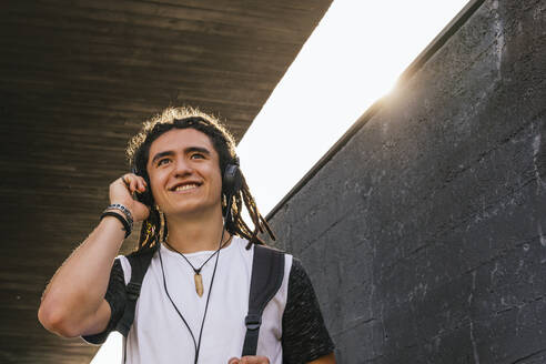 Lächelnder junger Mann, der wegschaut, während er über Kopfhörer an der Wand Musik hört - MGRF00260