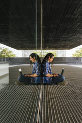 Young man using laptop while sitting by glass wall - MGRF00262