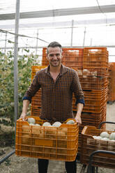 Smiling farmer carrying crate of melons at greenhouse - MPPF01741