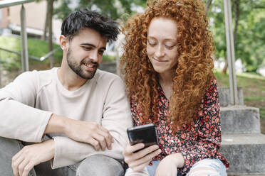 Lächelndes junges Paar benutzt Smartphone auf Stufen im Park - JCZF00761