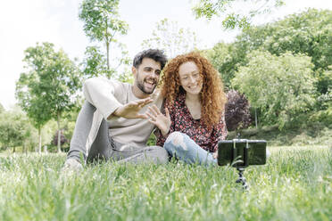 Happy young couple waving while vlogging in public park - JCZF00774