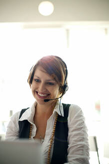 Smiling businesswoman wearing headset during video call on laptop in cafe - AJOF01429