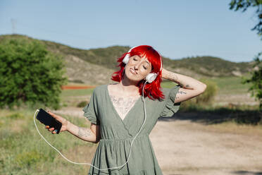 Young woman with hand in hair listening music on field during sunny day - MRRF01158