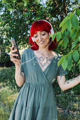 Smiling woman with smart phone listening music by tree - MRRF01160