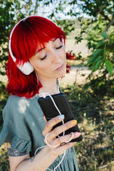 Schöne Frau mit geschlossenen Augen hält Handy beim Hören von Musik vor einem Baum - MRRF01161