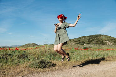 Junge Frau mit Mobiltelefon, die an einem sonnigen Tag durch ein Mohnfeld springt - MRRF01169