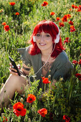 Smiling young woman with smart phone listening music on poppy field - MRRF01175