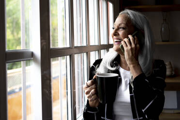 Glückliche Frau mit Kaffeetasse, die durch das Fenster schaut, während sie zu Hause mit ihrem Smartphone telefoniert - ERRF04914