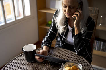 Frau mit digitalem Tablet, die am Tisch im Wohnzimmer sitzt und mit ihrem Mobiltelefon spricht - ERRF04922