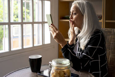 Ältere Frau küsst sich während eines Videoanrufs über ihr Smartphone zu Hause - ERRF04926