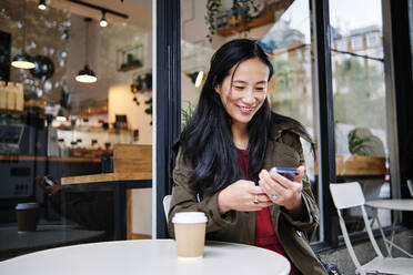 Smiling beautiful woman using mobile phone at sidewalk cafe - ASGF00361