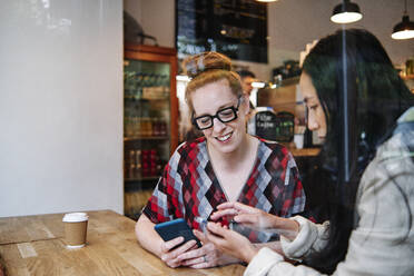 Woman showing smart phone to smiling female friends while sitting at table in cafe - ASGF00364