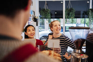 Smiling female friends looking away while standing in front of owner at cafe counter - ASGF00382