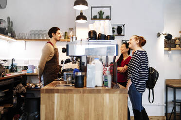 Lächelnde weibliche Kunden, die eine Hängelampe betrachten, während sie vor dem Besitzer eines Cafés stehen - ASGF00383