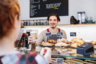 Lächelnder männlicher Besitzer bietet einer weiblichen Kundin in einem Café Kaffee an - ASGF00391