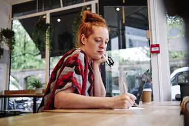 Woman with hand on chin writing on paper in cafe - ASGF00411