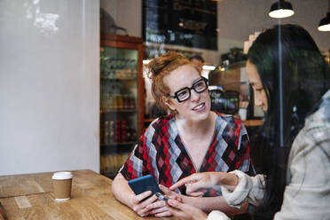 Frau mit Brille im Gespräch mit einer Freundin, die ein Mobiltelefon in einem Café benutzt - ASGF00415