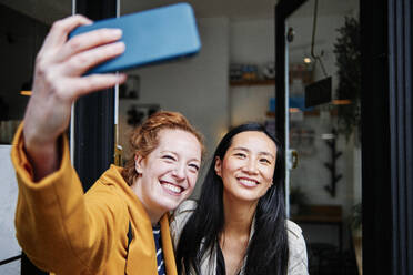 Lächelnde Frau nimmt Selfie durch Handy mit weiblichen Freund außerhalb Cafe - ASGF00423