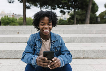 Afro woman using mobile phone while sitting on steps - XLGF01987