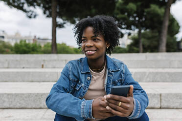 Smiling Afro woman holding mobile phone while sitting on steps - XLGF01989