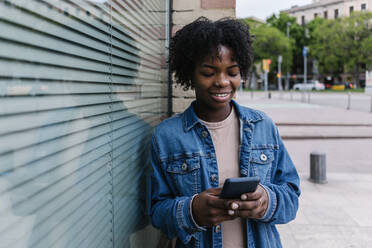 Afro-Frau, die ein Smartphone benutzt und sich an ein Fenster lehnt - XLGF02006