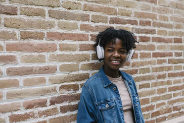 Smiling woman listening music through headphones while leaning on brick wall - XLGF02009