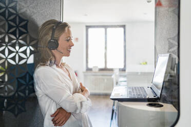 Smiling female customer service representative with arms crossed standing in front of laptop at office - DIGF15688