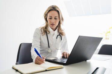 Eine Ärztin schreibt vor einem Laptop in einer Klinik an einem Tagebuch - DIGF15722