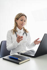 Ärztin gestikuliert während einer Videokonsultation über einen Laptop am Schreibtisch - DIGF15724