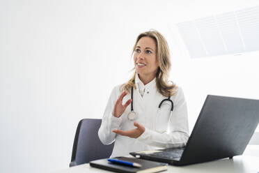 Ärztin schaut weg und gestikuliert während einer Videokonsultation am Schreibtisch - DIGF15725