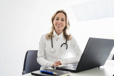 lächelnde Ärztin mit Stethoskop am Schreibtisch sitzend im Büro - DIGF15726