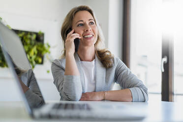 Smiling businesswoman looking away while talking on smart phone at desk - DIGF15743