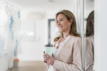 Smiling female entrepreneur with reusable coffee cup leaning on soundproof cabin at office - DIGF15750