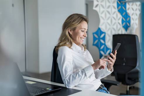 Lächelnde weibliche Fachkraft, die ein Smartphone am Schreibtisch im Büro benutzt - DIGF15753