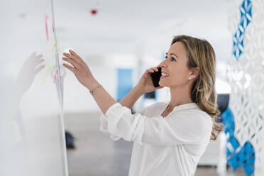 Smiling female professional talking on smart phone in front of whiteboard at office - DIGF15768