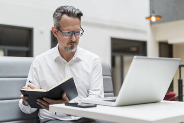 Männlicher Unternehmer mit Buch, der im Büro auf seinen Laptop schaut - DIGF15774
