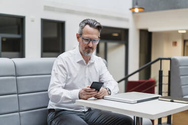 Männlicher Unternehmer, der ein Mobiltelefon benutzt, während er am Tisch im Büro sitzt - DIGF15776