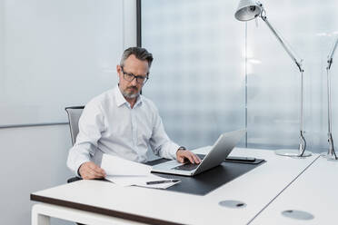 Männlicher Unternehmer mit Laptop, der im Büro sitzend Papier prüft - DIGF15822