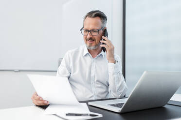 Geschäftsmann mit Dokument und Laptop, der im Büro mit einem Mobiltelefon spricht - DIGF15823