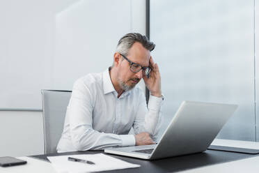 Geschäftsmann mit Kopf in der Hand sitzt mit Laptop am Schreibtisch - DIGF15826