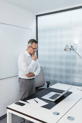 Thoughtful businessman with hand on chin looking at laptop in office - DIGF15831