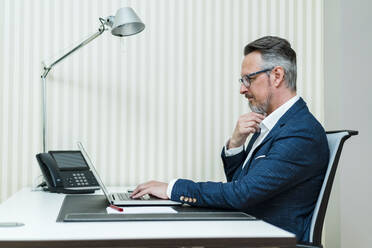 Männlicher Unternehmer mit Hand am Kinn, der einen Laptop am Schreibtisch benutzt - DIGF15844