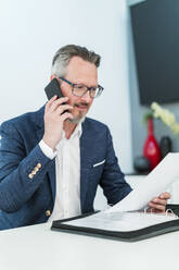 Männlicher Unternehmer prüft ein Dokument, während er im Büro mit dem Handy telefoniert - DIGF15855