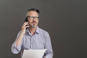 Männlicher Unternehmer mit Dokument, der mit seinem Smartphone vor einer grauen Wand spricht - DIGF15858