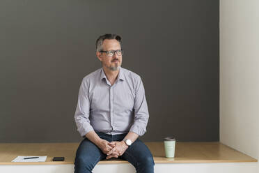 Mature businessman sitting by disposable cup and mobile phone in office - DIGF15859